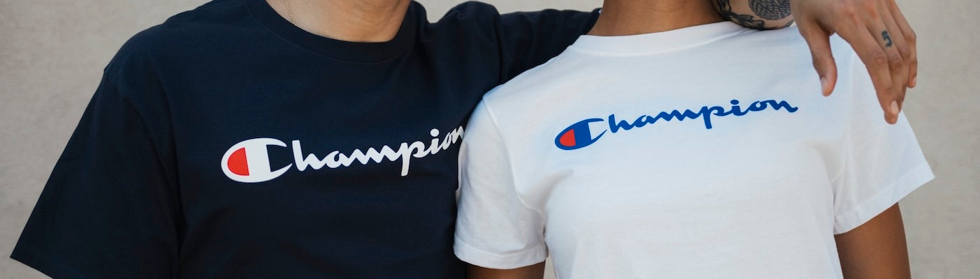 Two people wearing black and white Champion t-shirts stand side by side with one persons arm around the other, showing the brands logo clearly on both shirts.