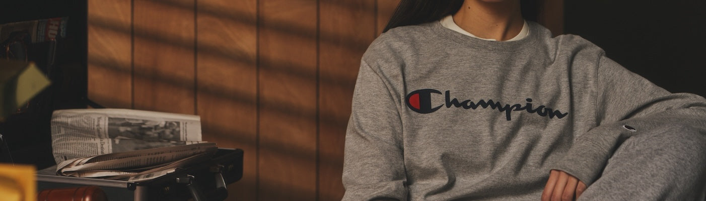 A person wearing a grey Champion sweatshirt and matching pants sits indoors next to a table with newspapers and magazines. Wood paneling and sunlight create a warm background.