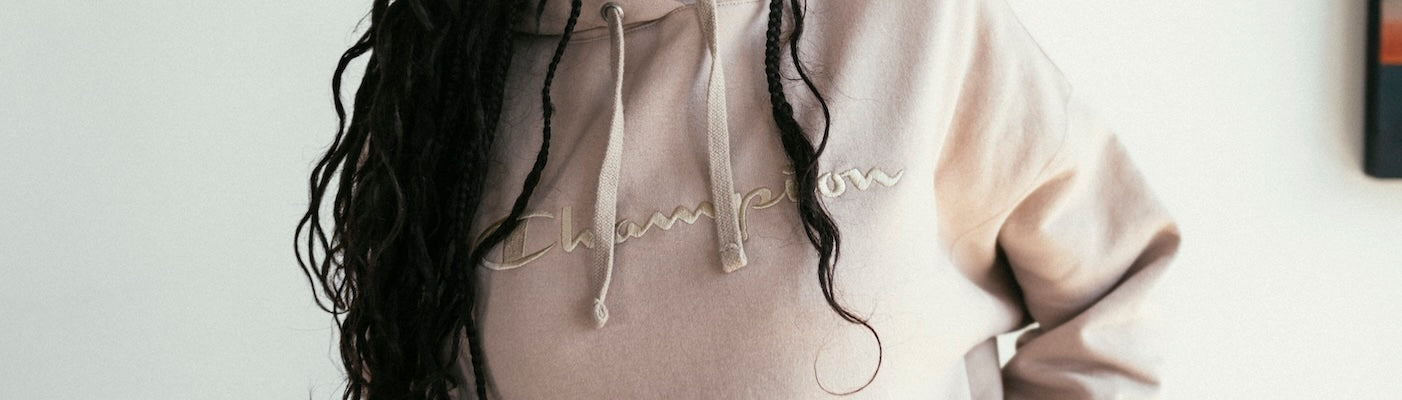 Person with long braids wearing a light beige Champion hoodie, shown from the shoulders to the waist against a plain background.