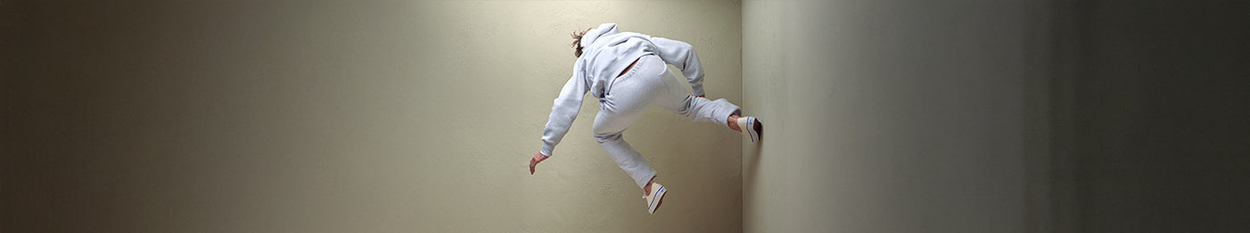 A person in a light gray tracksuit appears to be running or climbing sideways on a beige wall in a narrow, dimly lit room.