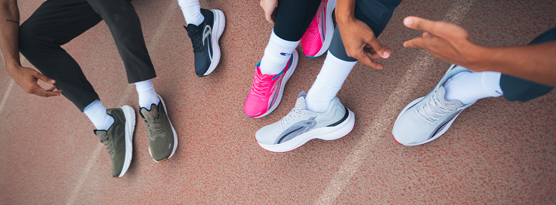 Four people sit on a track, showing only their legs and feet. Each wears white socks and different colored running shoes: olive green, black, pink, and light gray.