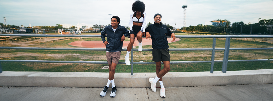 Three young adults in athletic wear relax by a railing next to a sports track; two lean against the rail while one sits on top, all appearing happy and casual outdoors.