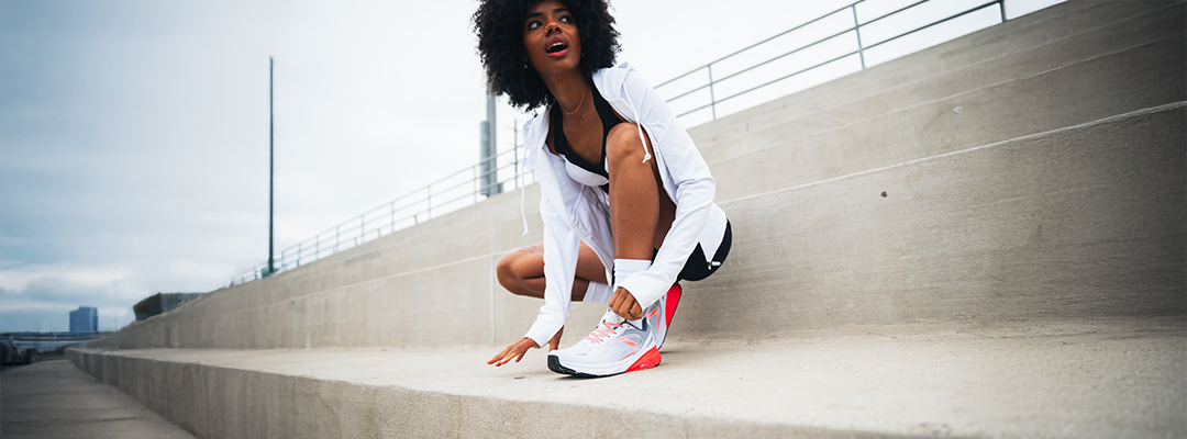A woman in athletic wear crouches on outdoor concrete steps, looking up and smiling, with one hand on her knee and the other adjusting her white and pink sneaker.
