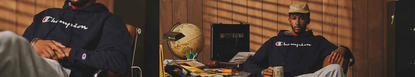 A man wearing a navy Champion hoodie and light pants sits at a desk with a globe, folders, and stationery, holding a mug. Sunlight casts shadows from blinds on the wood-paneled wall.