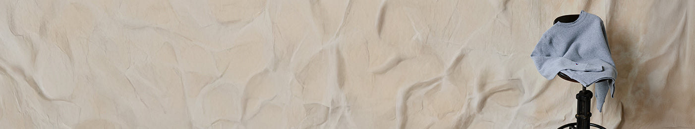 A light blue long-sleeve shirt draped over the back of a black chair, set against a beige, textured wall with a crumpled appearance.