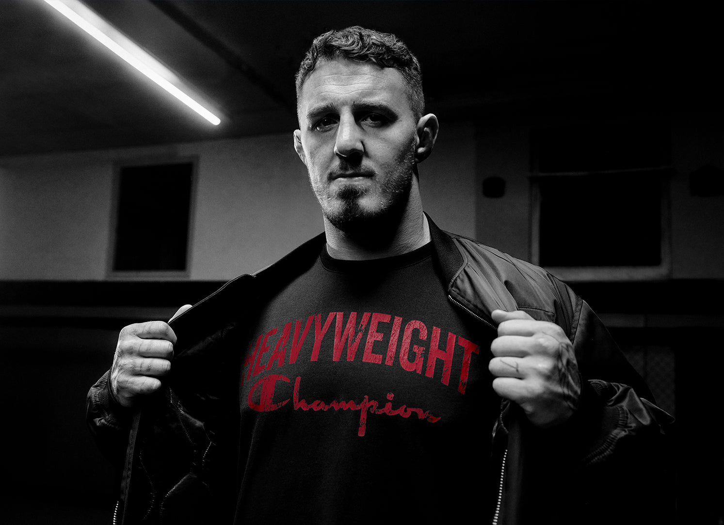 A man in a dark jacket opens it to reveal a black shirt with red text that reads HEAVYWEIGHT Champions. He stands in a dimly lit room, looking confidently at the camera.