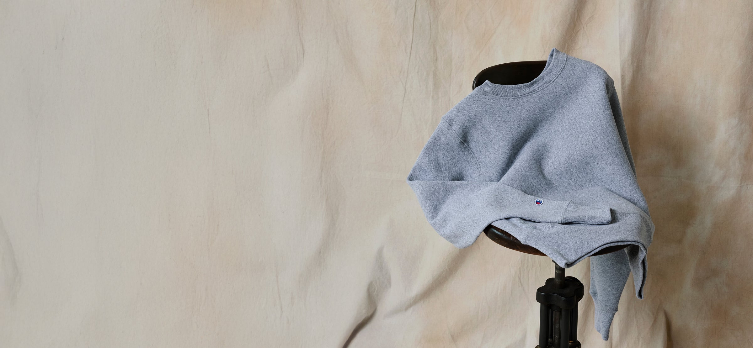 A light gray crewneck sweater draped over the back of a dark stool, set against a beige, slightly wrinkled fabric background.