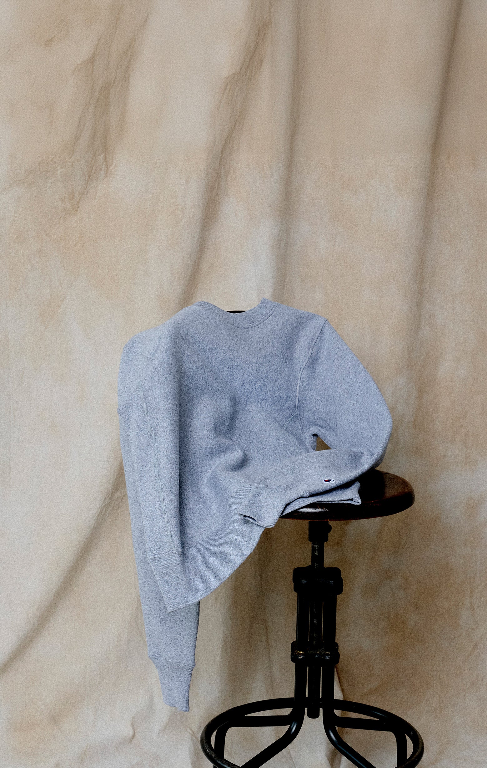 A light gray sweatshirt draped over a black adjustable stool, set against a beige, slightly wrinkled fabric backdrop.