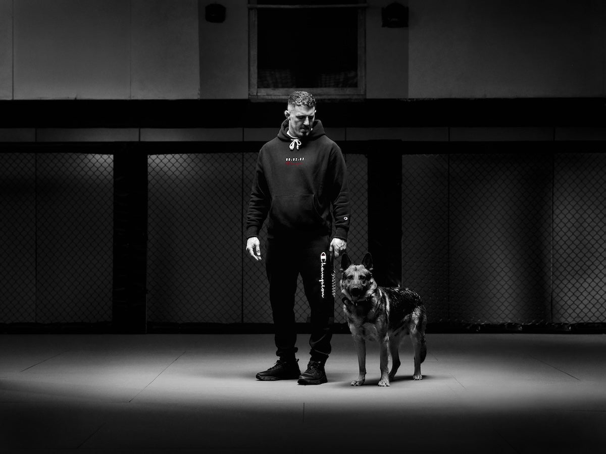A man in dark athletic clothing stands in a spotlight next to a German Shepherd dog inside an empty gym with chain-link fencing in the background. The photo is black and white.