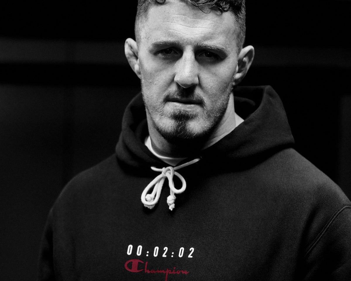A man with short hair and a beard wears a hoodie with the word Champion and numbers 00:02:02 on it, looking seriously at the camera in a monochrome setting.