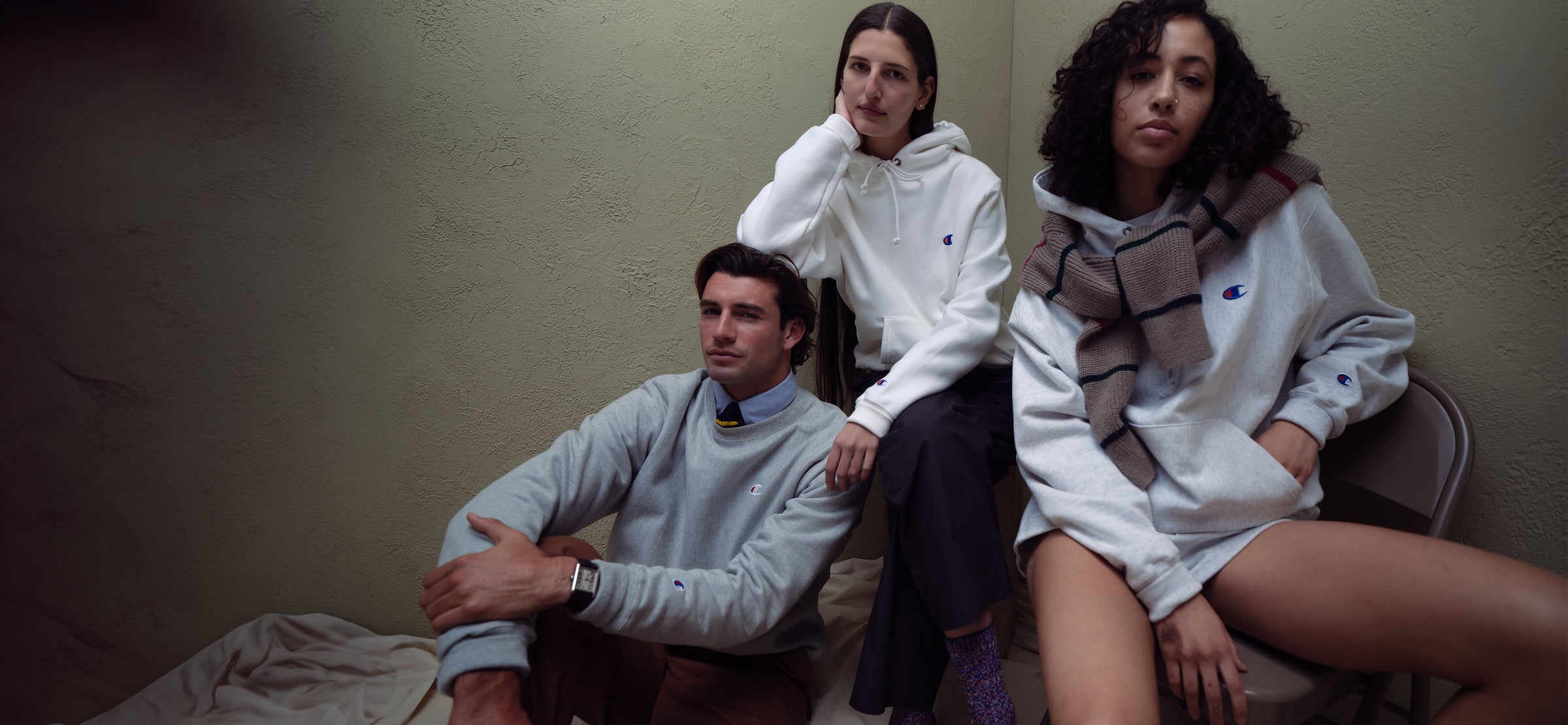 Three people sit indoors wearing Champion hoodies and sweatshirts. One person leans against the wall, one sits on the floor, and another sits on a chair with a plaid scarf. The setting is casual and relaxed.