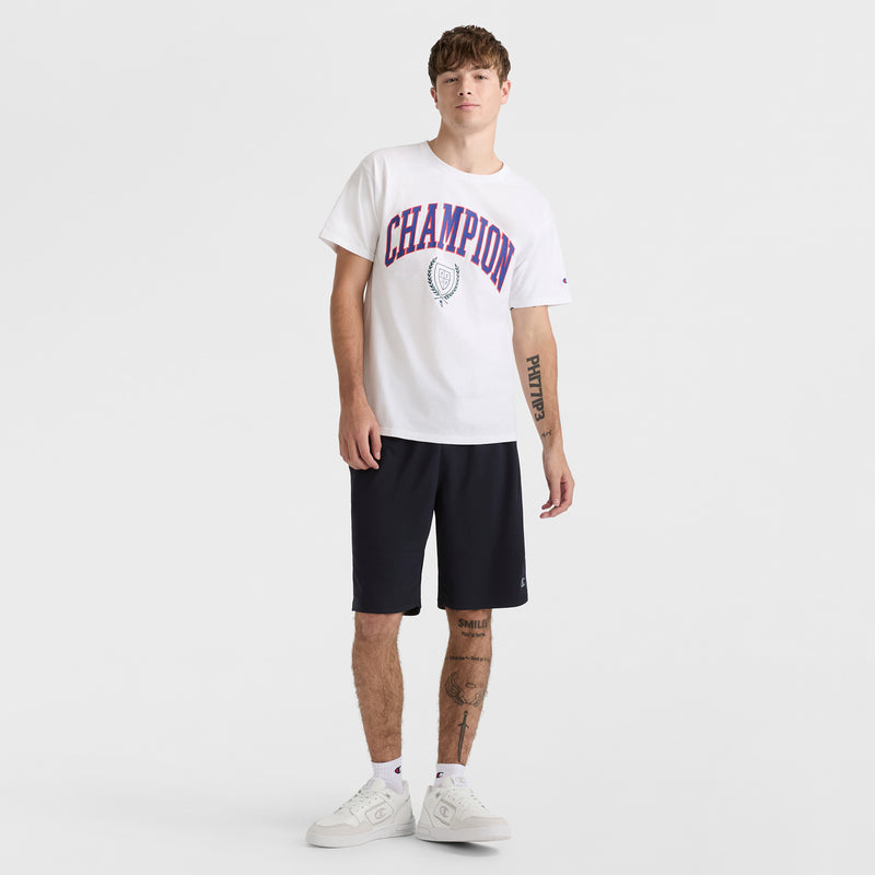 A young man with tattoos stands casually against a white backdrop wearing a white Champion tee, 10" Moisture Wicking Sport Shorts by Champion in black, and white sneakers, with one hand partly in his pocket.