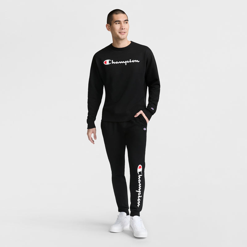 A man stands against a white background wearing a black Champion Powerblend Crewneck Sweatshirt, Classic Script Logo, and matching joggers with white sneakers. The Champion logo is visible on both the sweatshirt and pants.