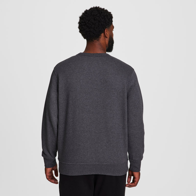 A man with short curly hair faces away from the camera, wearing a dark gray Champion Powerblend Crewneck Sweatshirt, C Logo - Big & Tall, and black pants against a plain white background.