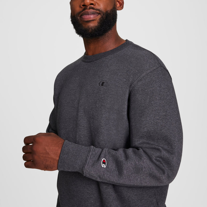 A man in a dark gray Champion Powerblend Crewneck Sweatshirt, C Logo - Big & Tall, stands against a plain background. The fleece pullover displays the Champion logo on the chest and sleeve; only his lower face is visible.