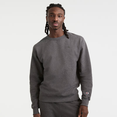 A man with long braided hair wears a dark gray Champion Powerblend Crewneck Sweatshirt, C Logo, paired with matching sweatpants, standing against a plain white background.