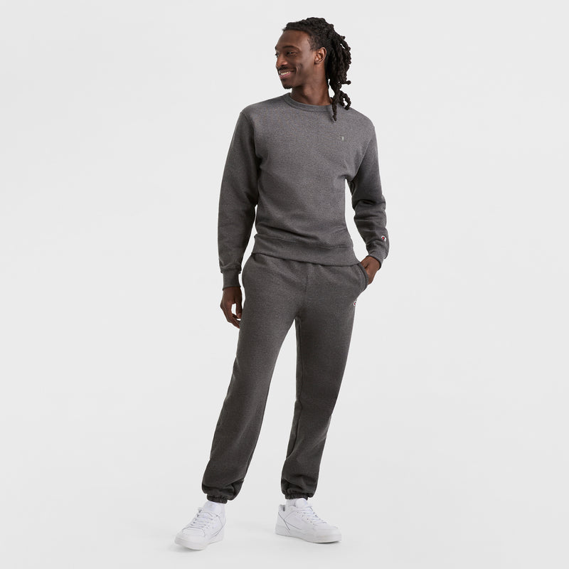 Wearing a matching dark gray Champion Powerblend Crewneck Sweatshirt with the C Logo and sweatpants, paired with white sneakers, a person stands against a plain white background, looking to the side and smiling.