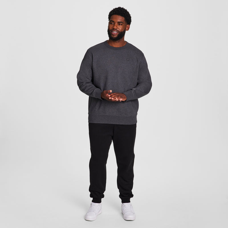 A man smiles against a white background, wearing the Champion Powerblend Crewneck Sweatshirt, C Logo - Big & Tall in dark gray, paired with black sweatpants and white sneakers. He clasps his hands, highlighting the cozy style.