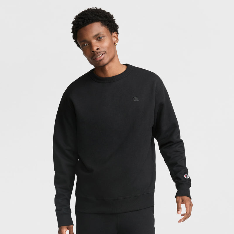 A man with short curly hair wears a black Champion Powerblend Crewneck Sweatshirt, C Logo, with matching pants, standing against a plain white background.
