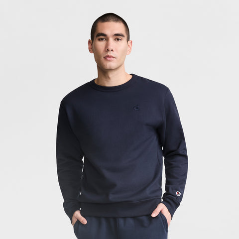 A man wears a Champion Powerblend Crewneck Sweatshirt, C Logo, paired with navy blue pants, standing with hands in his pockets against a plain white background.