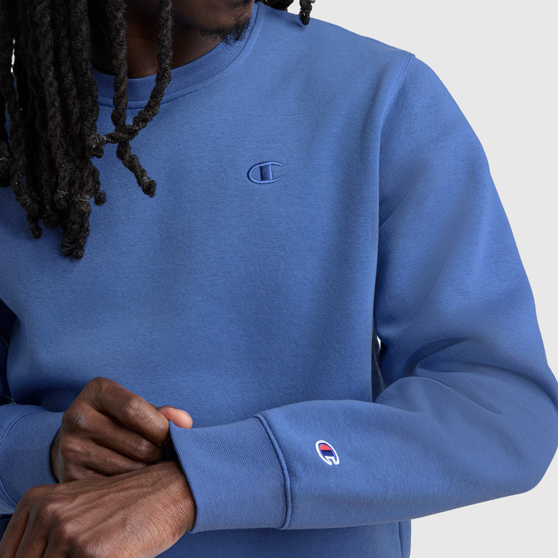 A person with long, twisted hair (face unseen) adjusts the sleeve of a blue Champion Powerblend Crewneck Sweatshirt, C Logo. The Champion emblem appears on both the chest and sleeve.