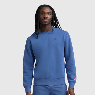 A man with long braided hair wears a blue Champion Powerblend Crewneck Sweatshirt, C Logo, and matching pants, standing against a plain light gray background.