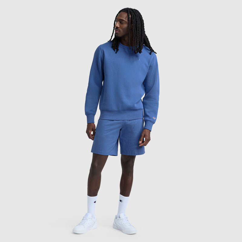 A man with long braided hair wears a blue Champion Powerblend Crewneck Sweatshirt, C Logo, and matching shorts with white crew socks and sneakers, standing against a plain light gray background.