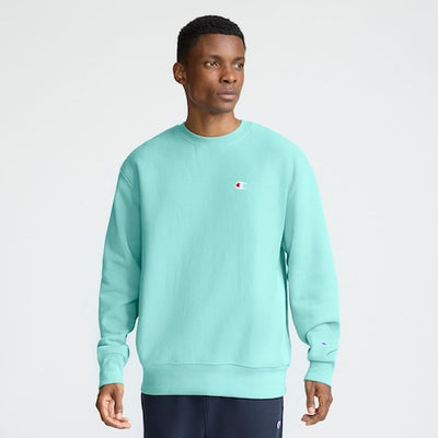 A man wearing a light turquoise Champion sweatshirt and dark pants stands against a plain light background, looking slightly to the side.