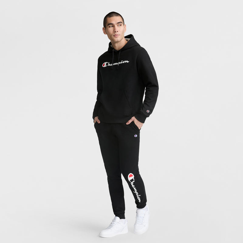 A man stands against a white background wearing a black Champion Powerblend Hoodie with a full script logo and matching sweatpants, plus white sneakers.