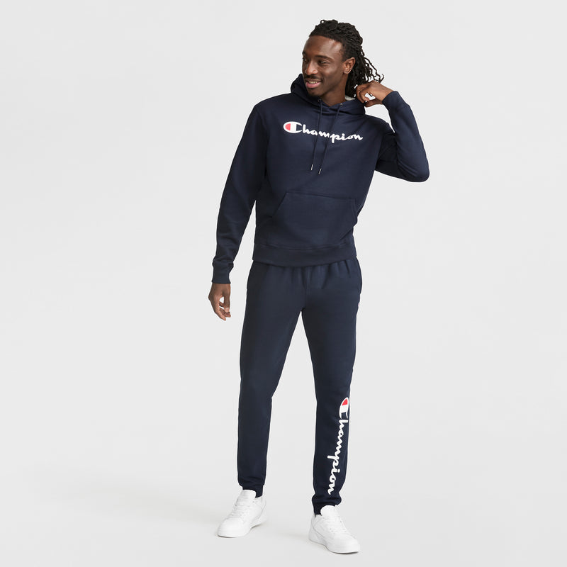 A man wearing a navy blue Champion Powerblend Hoodie with matching sweatpants and white sneakers stands smiling and looking to the side against a plain white background.