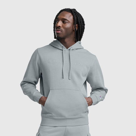 A man with shoulder-length dreadlocks wears a light gray Champion Powerblend Hoodie with a tonal script logo and matching sweatpants, standing hands-in-pocket against a plain light gray background.