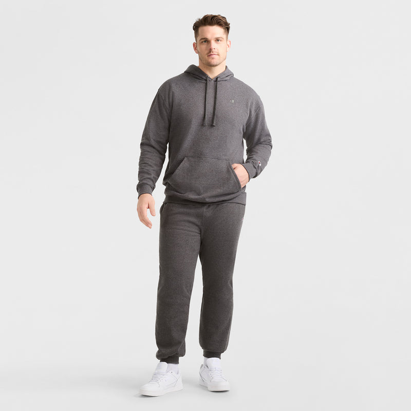 A man stands against a plain white background wearing the Champion Powerblend Hoodie, C Logo - Big & Tall in dark gray with matching sweatpants and white sneakers. One hand is in his pocket while the other rests by his side.
