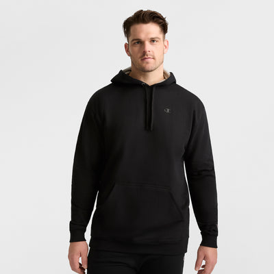 A man faces forward in a plain black Champion Powerblend Hoodie, C Logo - Big & Tall, with a small logo on the left chest and black pants, against a light gray background.