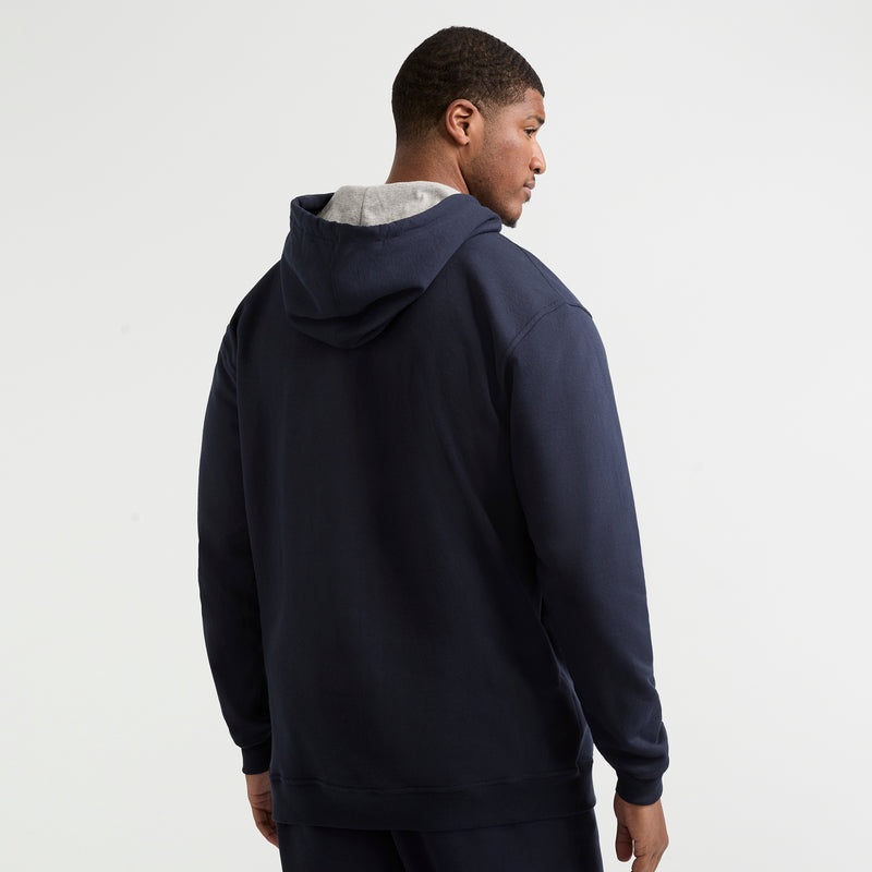 A man faces away from the camera, wearing a Champion Powerblend Hoodie, C Logo – Big & Tall in dark blue with the hood down. The background is plain white.