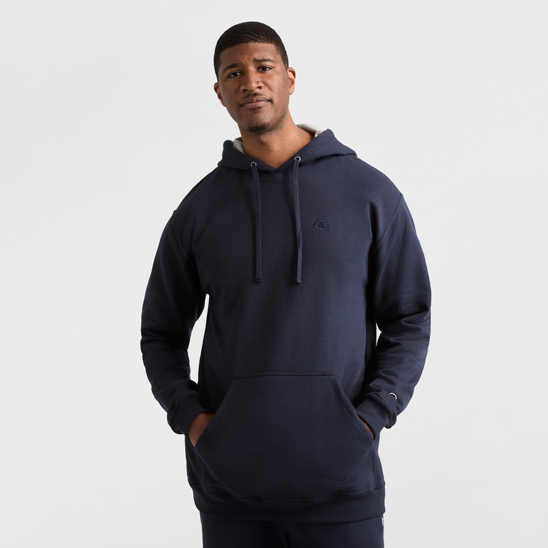 A man stands against a plain light background, wearing the Champion Powerblend Hoodie, C Logo - Big & Tall in navy blue with a front pocket and drawstrings. His hands are in the pocket, and he has a neutral expression.