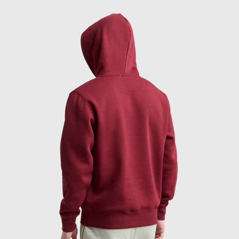 A person wearing a red Champion Powerblend Hoodie, C Logo, with the hood up and facing away from the camera. The background is plain light gray, and their hands rest relaxed by their sides.