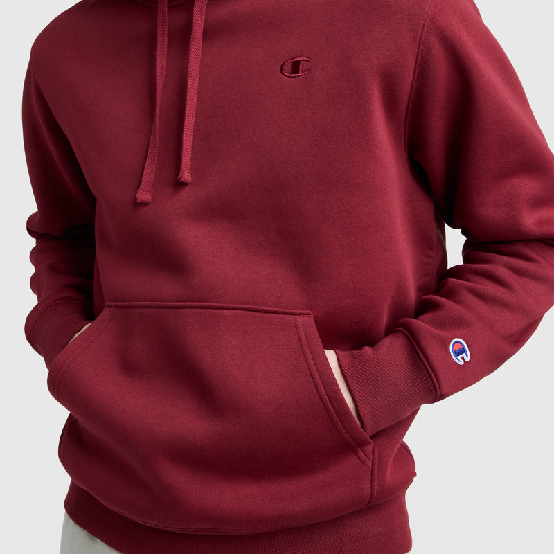 A person wears the Champion Powerblend Hoodie, C Logo, in maroon with hands in the front pocket. The Champion logo appears on the chest and left sleeve. The men's hoodie is shown against a plain light gray background.