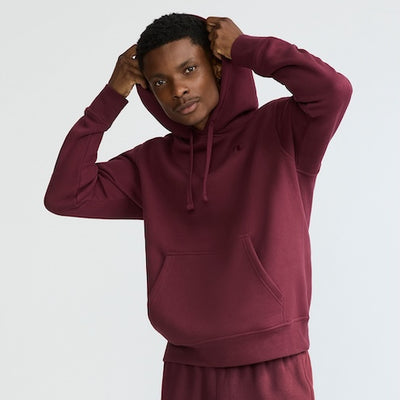 A person wearing a maroon hoodie and matching pants adjusts the hood, looking confidently at the camera against a plain light background.