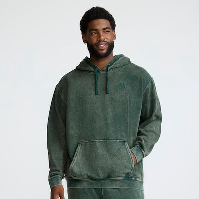 A man wearing a green, acid-wash hoodie with a front pocket and matching drawstrings stands against a plain light background, looking slightly to the side and smiling.