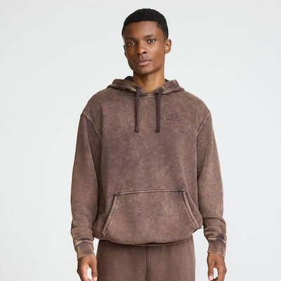A person stands facing forward, wearing a brown, faded, oversized hoodie with a front pocket and matching pants, against a plain light background.