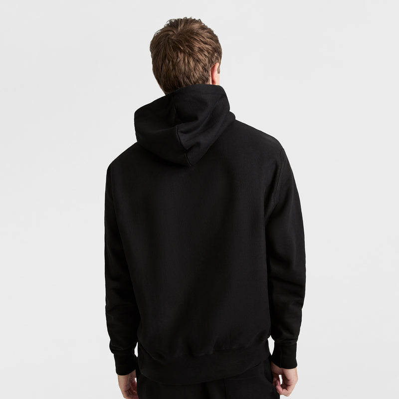 A person with short brown hair faces away from the camera, wearing a plain black Champion Reverse Weave Hoodie with the C Logo and the hood down, against a plain white background.