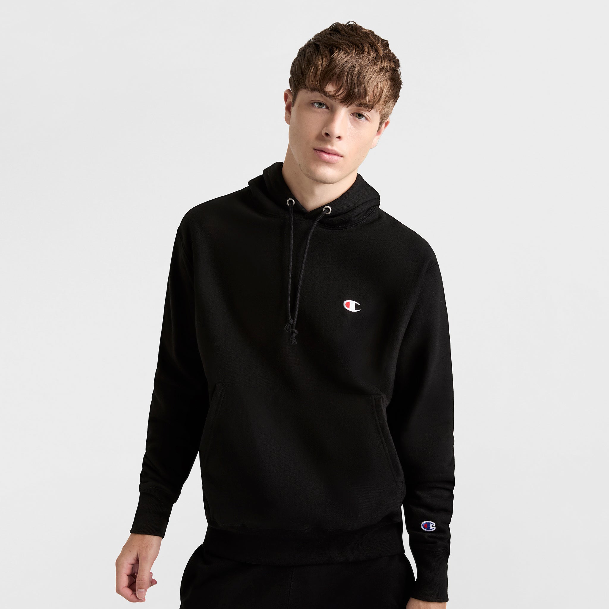 A young man with short brown hair wears a black Champion Reverse Weave Hoodie, C Logo—a heavyweight pullover with small chest and sleeve logos—standing against a plain light background.