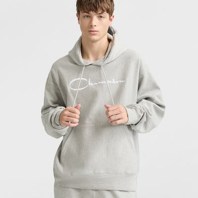 A young person wearing a light gray hooded sweatshirt with Charmism written in white script on the front, standing against a plain light background and looking at the camera.