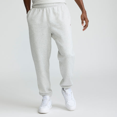 A person wearing Champion Powerblend Sweatpants, Cinched Hem, C Logo - Big & Tall in light gray and white sneakers stands with hands in pockets against a plain white background. The upper body is mostly out of frame.
