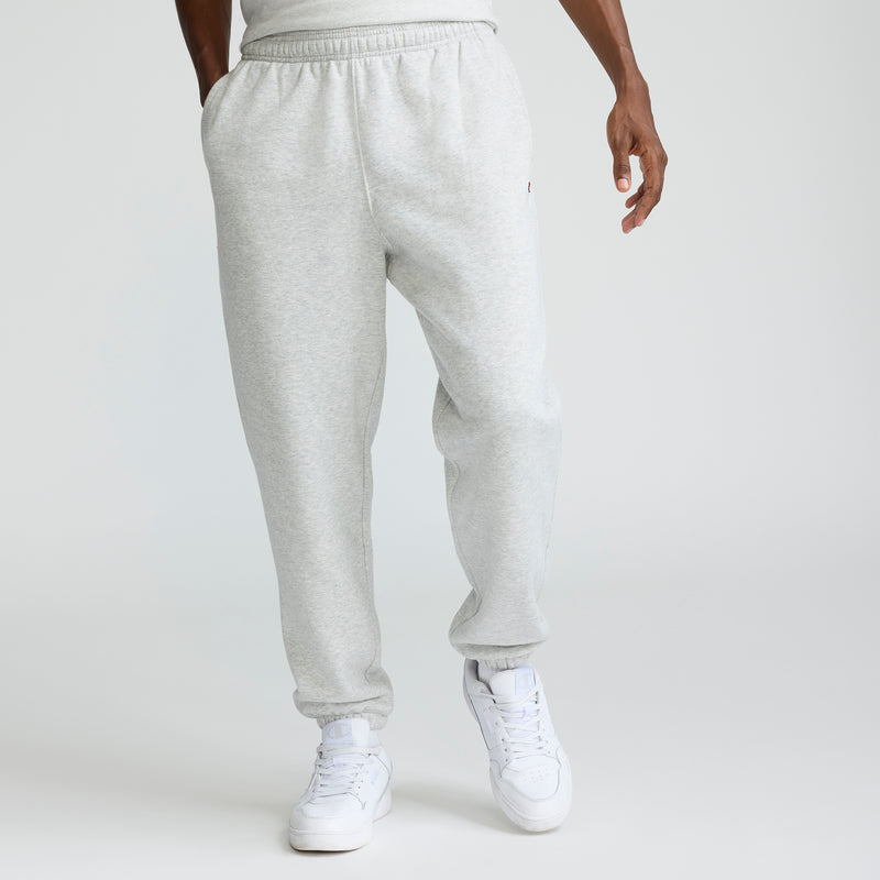 A person wearing Champion Powerblend Sweatpants, Cinched Hem, C Logo - Big & Tall in light gray and white sneakers stands with hands in pockets against a plain white background. The upper body is mostly out of frame.