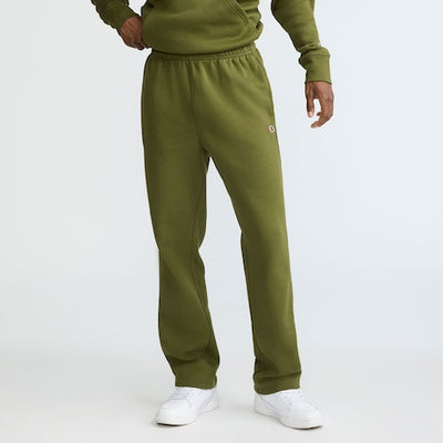 A person wearing an olive green sweatshirt and matching sweatpants stands with one hand on their hip. They are also wearing white sneakers. The background is plain and light-colored.
