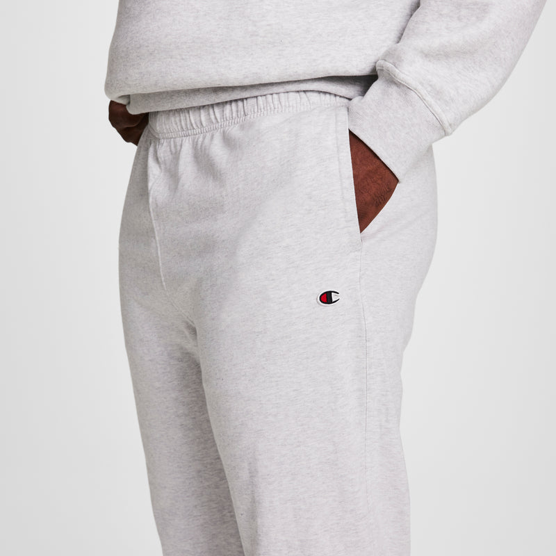 A person wears Champion Lightweight Lounge Pants, Open Hem, C Logo - Big & Tall in light gray with a matching sweatshirt, standing with one hand in their pocket. The Champion logo is visible on the pants.