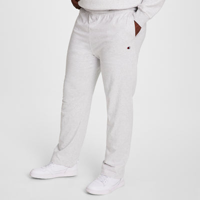 A person wears Champion Big & Tall Lightweight Lounge Pants, Open Hem, C Logo in light gray with hands in pockets and white sneakers. Only the lower body is shown against a plain white background. Soft jersey knit fabric provides extra comfort.