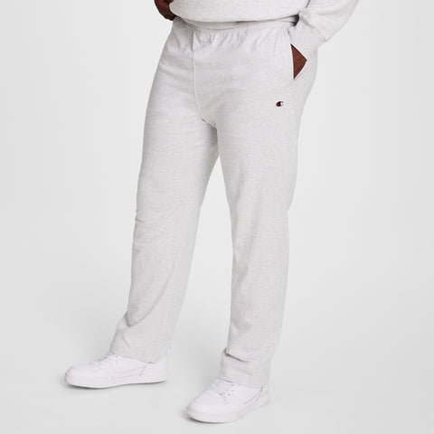 A person wears Champion Big & Tall Lightweight Lounge Pants, Open Hem, C Logo in light gray with hands in pockets and white sneakers. Only the lower body is shown against a plain white background. Soft jersey knit fabric provides extra comfort.