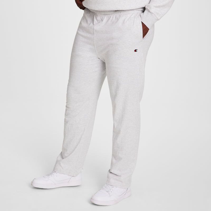 A person wears Champion Big & Tall Lightweight Lounge Pants, Open Hem, C Logo in light gray with hands in pockets and white sneakers. Only the lower body is shown against a plain white background. Soft jersey knit fabric provides extra comfort.